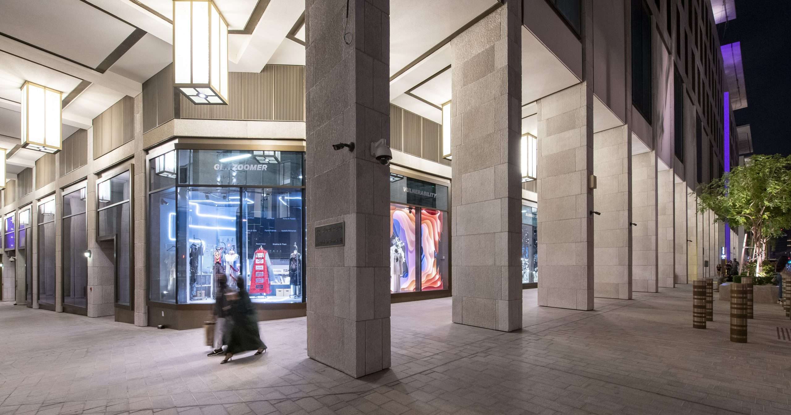 A modern building with large glass windows displaying clothing. A person walks by on a sidewalk in Msheireb Downtown.